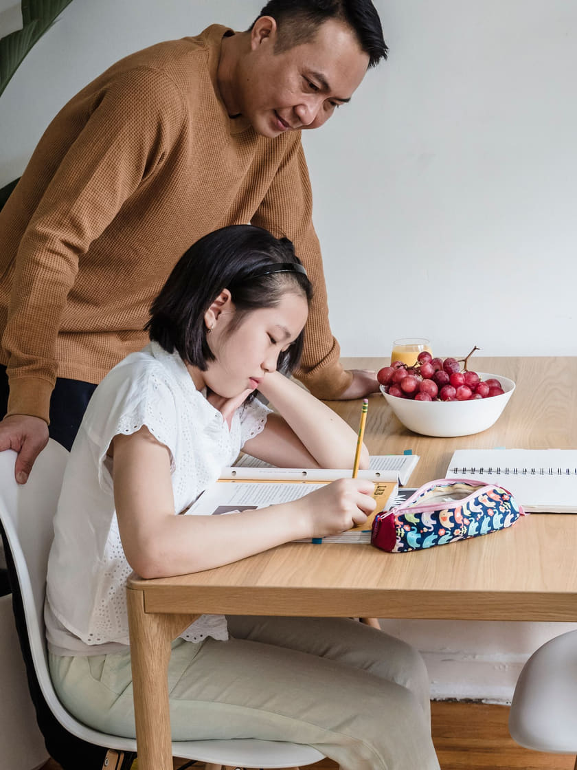 A child working through a math problem at a kitchen table while a parent looks on