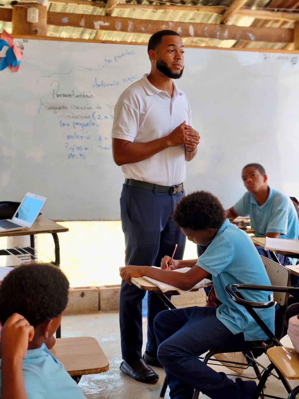 A teacher addressing students in a classroom while a student writes in a notebook